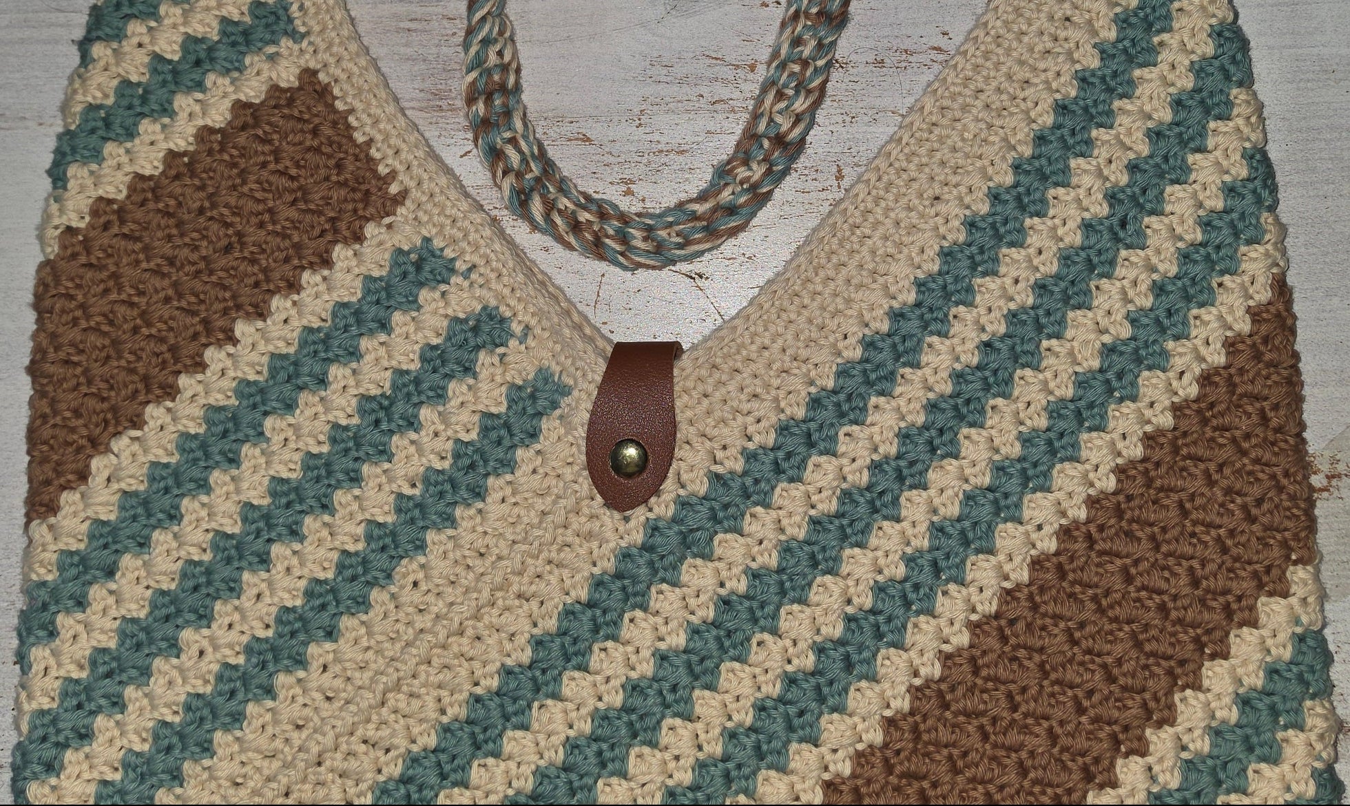 Crocheted shoulder bag with diagonal stripes in beige, brown, and teal, featuring braided rope handles and a small brown leather accent, displayed on a distressed white wooden surface.